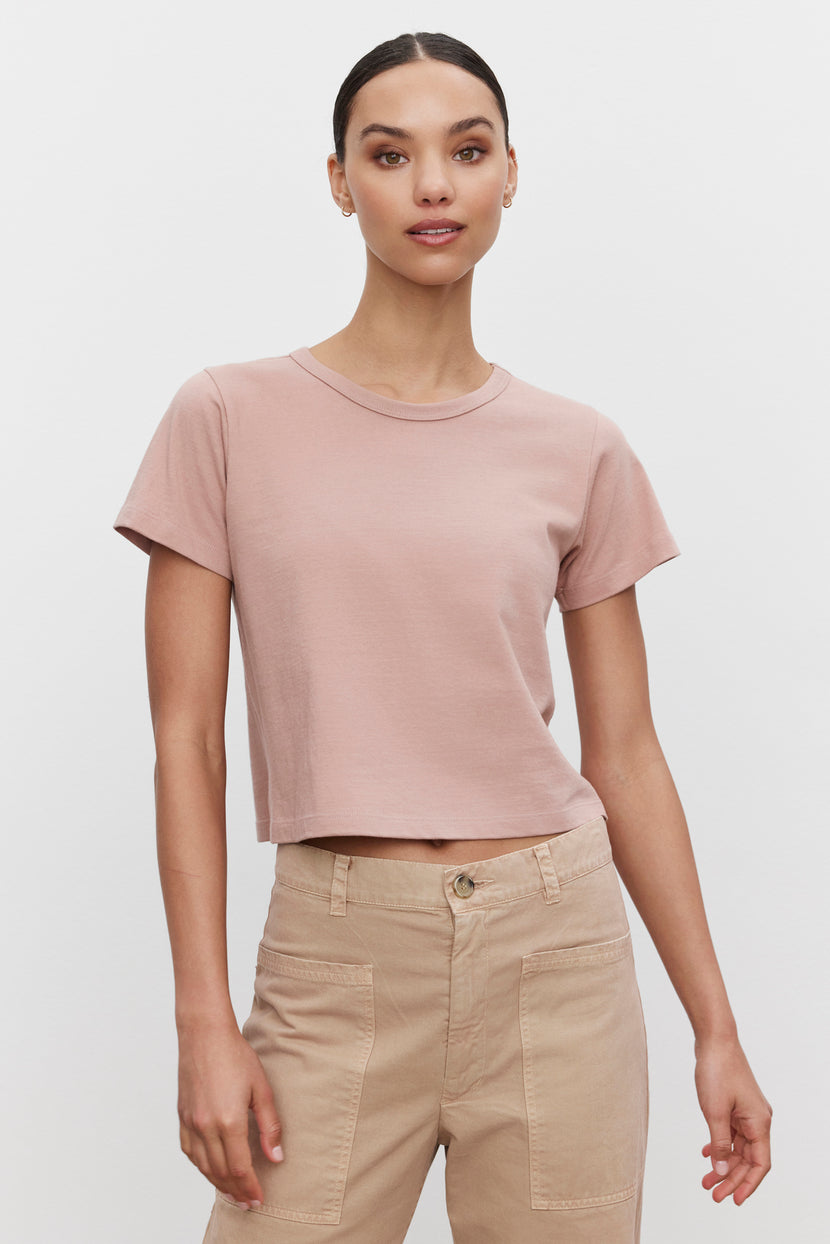 A person is standing and wearing the TINSLEY TEE, a light pink cropped tee in soft cotton by Velvet by Graham & Spencer, paired with beige high-waisted pants. They are looking at the camera against a plain white background, exuding a vintage feel.