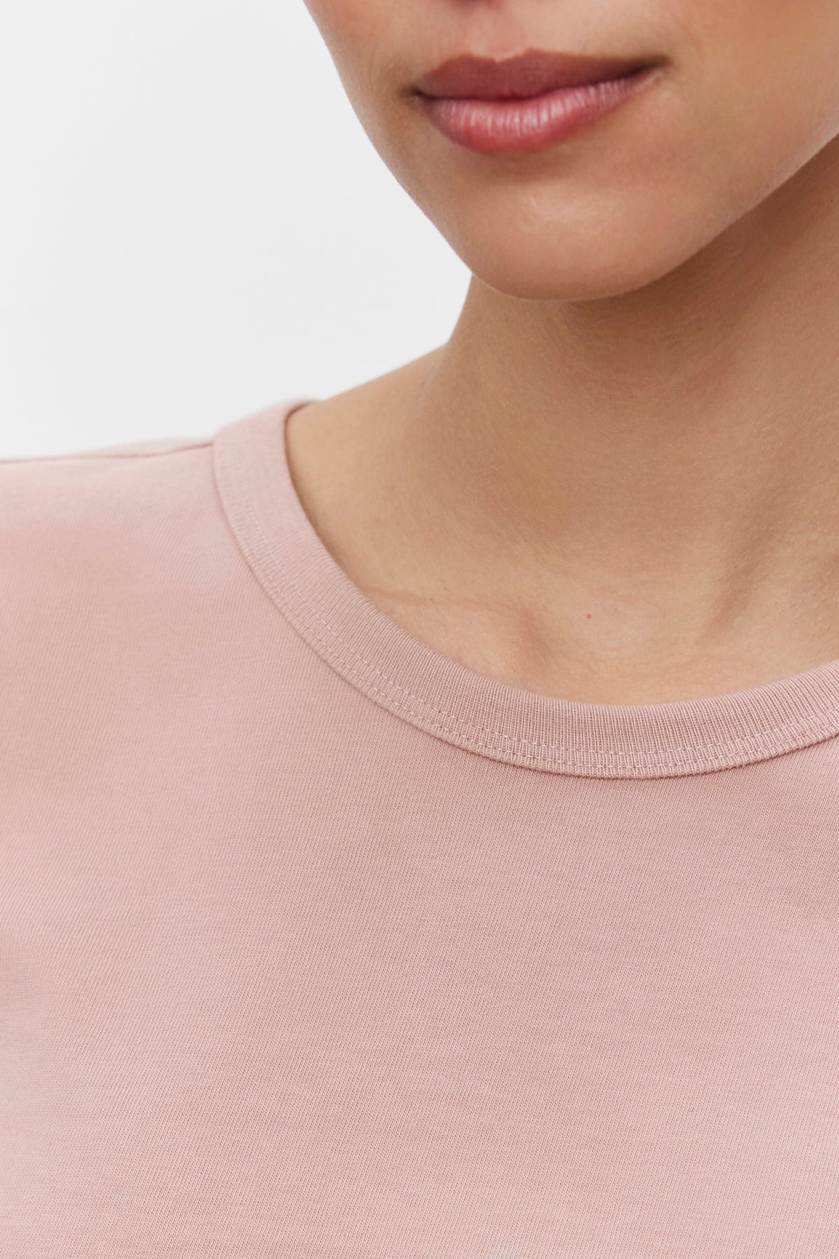   Close-up of a person wearing the TINSLEY TEE, a light pink, soft cotton t-shirt by Velvet by Graham & Spencer. The image focuses on the lower half of their face and the neckline of the shirt against a plain white background. 