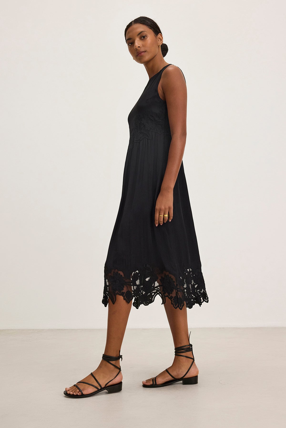   A person wears a BIANCA DRESS by Velvet by Graham & Spencer—a sleeveless black dress with a lace hem—and pairs it with black strappy sandals, standing against a plain white background. 