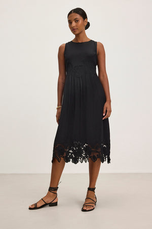 A person is wearing the BIANCA DRESS, a sleeveless black dress with floral lace at the hem by Velvet by Graham & Spencer, standing against a plain background.