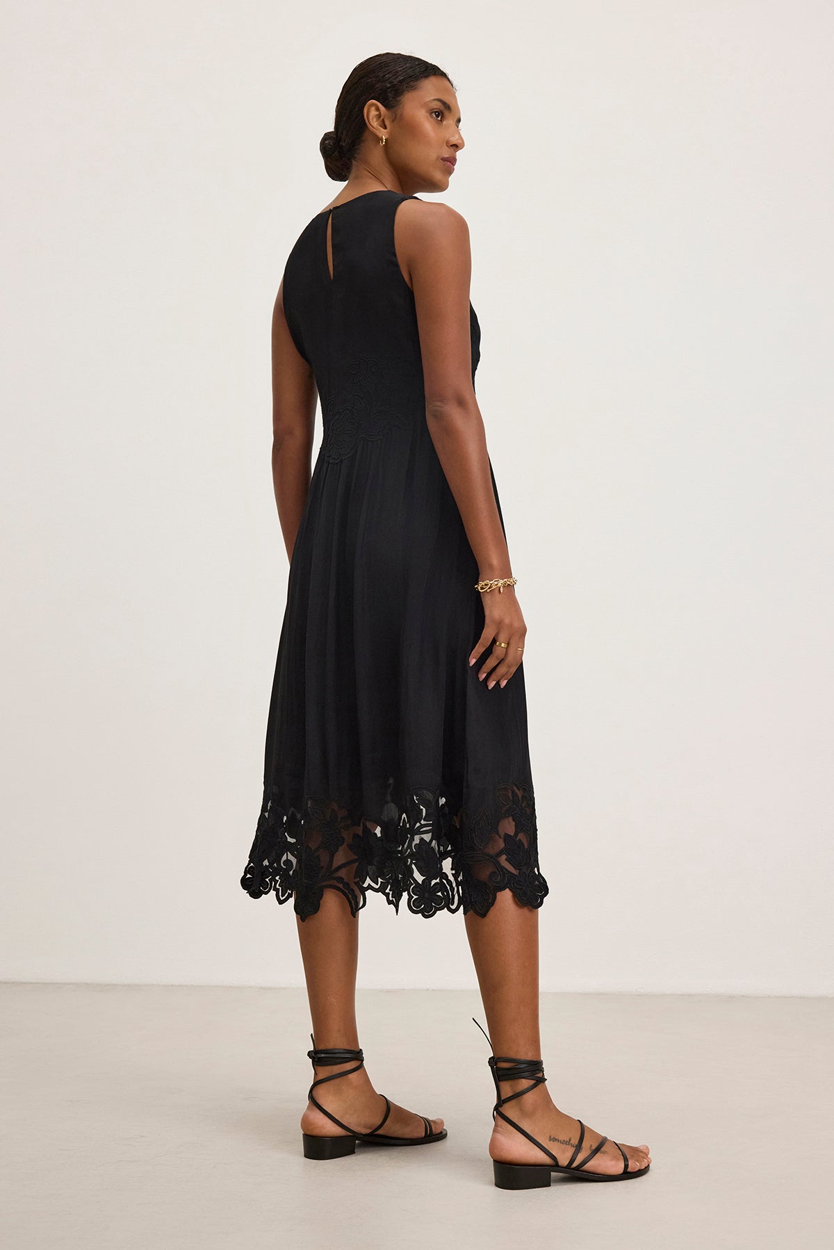   A woman in the BIANCA DRESS by Velvet by Graham & Spencer, featuring a sleeveless design and lace hem, is standing and looking over her shoulder. She accessorizes with strappy sandals and a bracelet. 