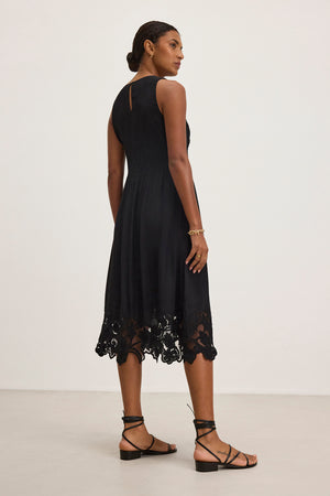 A woman in the BIANCA DRESS by Velvet by Graham & Spencer, featuring a sleeveless design and lace hem, is standing and looking over her shoulder. She accessorizes with strappy sandals and a bracelet.