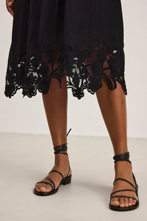 A model showcases the BIANCA DRESS by Velvet by Graham & Spencer, featuring a black skirt with floral embroidery, paired with black strappy sandals on a light-colored floor.