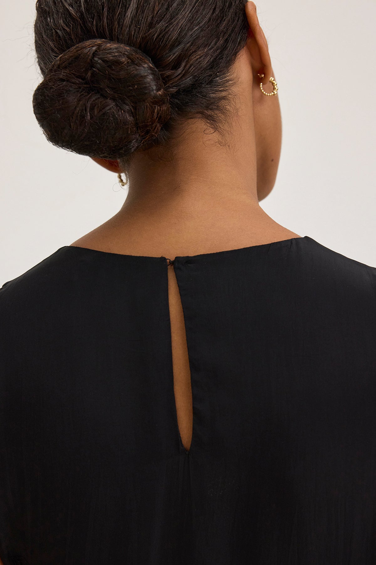   A person seen from behind wears the BIANCA DRESS by Velvet by Graham & Spencer, featuring hair in a bun, a black top with a small back slit, and a gold ear cuff. 