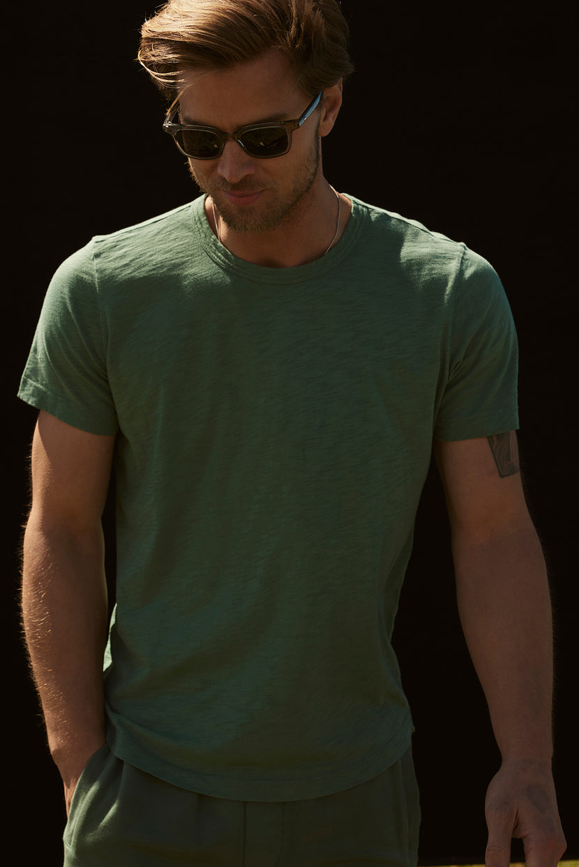 A man with sunglasses, wearing a stylish AMARO TEE in green by Velvet by Graham & Spencer, looks down with his hands in his pockets against a black background.