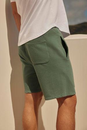 Man in a white t-shirt and Velvet by Graham & Spencer Beckett shorts standing beside a white railing, with a scenic background partially visible.