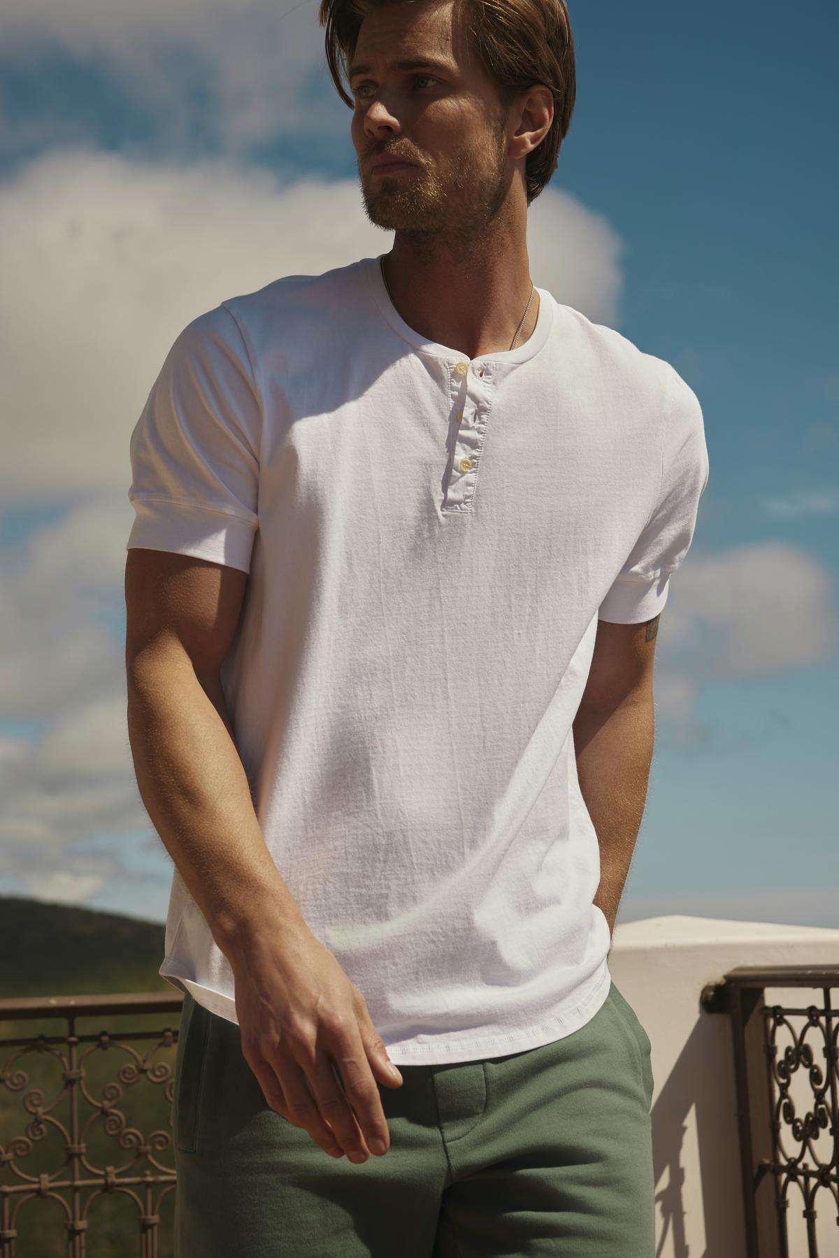 A man in a white Velvet by Graham & Spencer DEON HENLEY shirt with scooped hem and green trousers stands on a balcony, looking to the side with a backdrop of hills and sky.-36753582817473