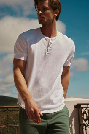 A man in a white Velvet by Graham & Spencer DEON HENLEY shirt with scooped hem and green trousers stands on a balcony, looking to the side with a backdrop of hills and sky.