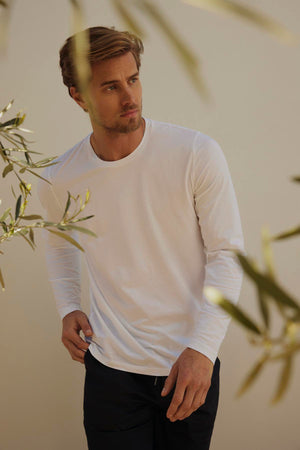 A man with short hair and a beard, wearing a long-sleeve white KAI TEE by Velvet by Graham & Spencer with a modern curved hemline and dark pants, is standing outdoors amidst some greenery.