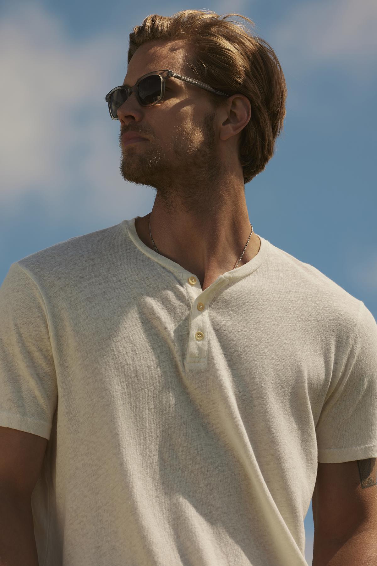   Man with blonde hair wearing sunglasses and a Velvet by Graham & Spencer's LIONEL HENLEY polo shirt looks up against a clear sky backdrop. 