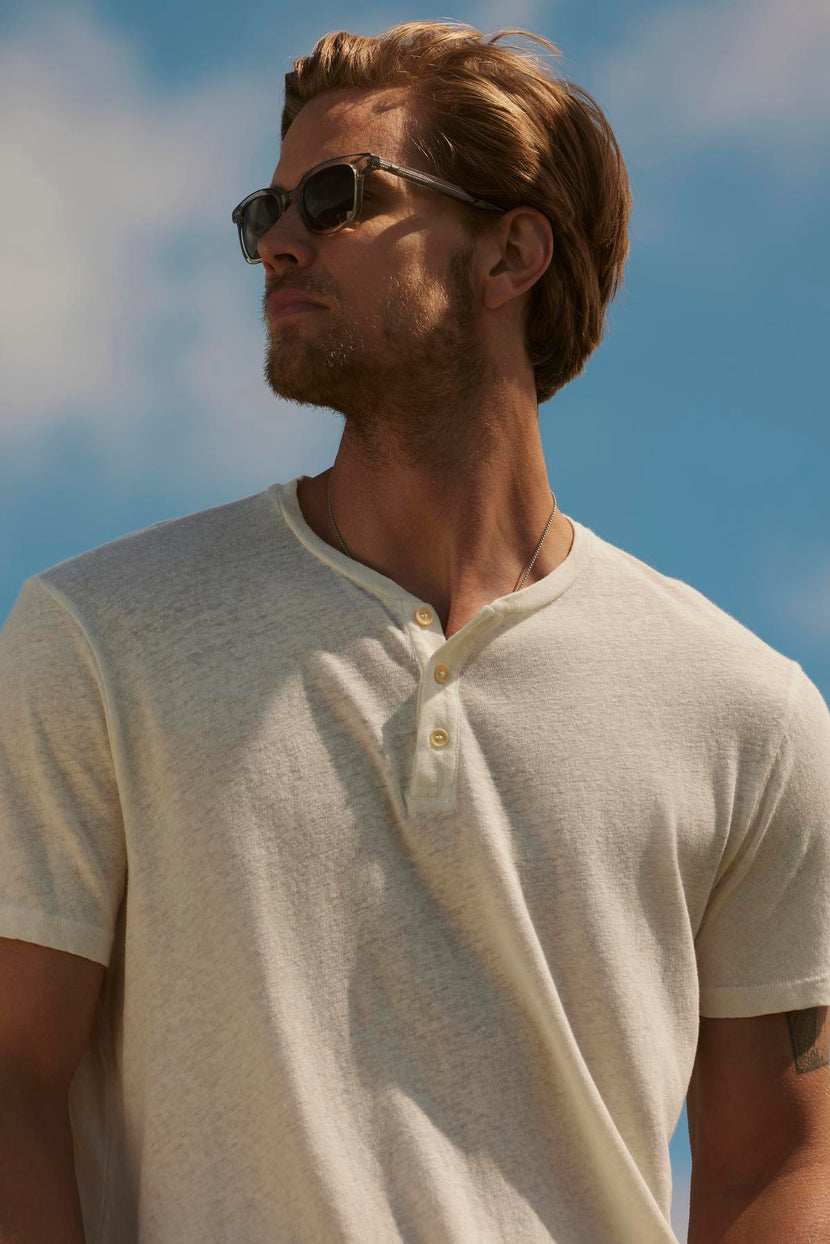 Man with blonde hair wearing sunglasses and a Velvet by Graham & Spencer's LIONEL HENLEY polo shirt looks up against a clear sky backdrop.