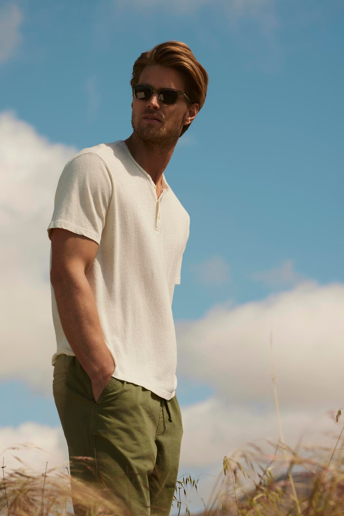 A man in a white Velvet by Graham & Spencer Lionel Henley shirt and green pants wearing sunglasses stands in a field, looking to the side under a cloudy sky.-36753588617409