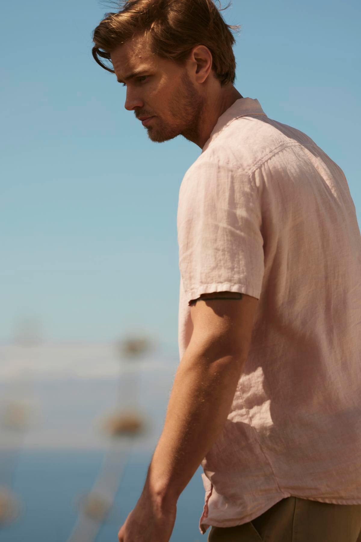   A man with light brown hair wearing a Velvet by Graham & Spencer MACKIE LINEN BUTTON-UP SHIRT gazes thoughtfully with a clear blue sky and blurry maritime background. 