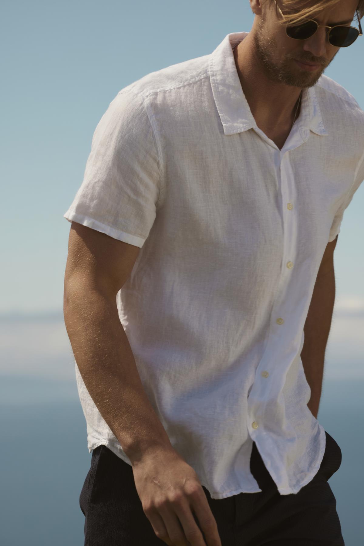   A man in a white relaxed fit Velvet by Graham & Spencer MACKIE LINEN BUTTON-UP SHIRT and sunglasses stands by the sea, a clear sky in the background. 