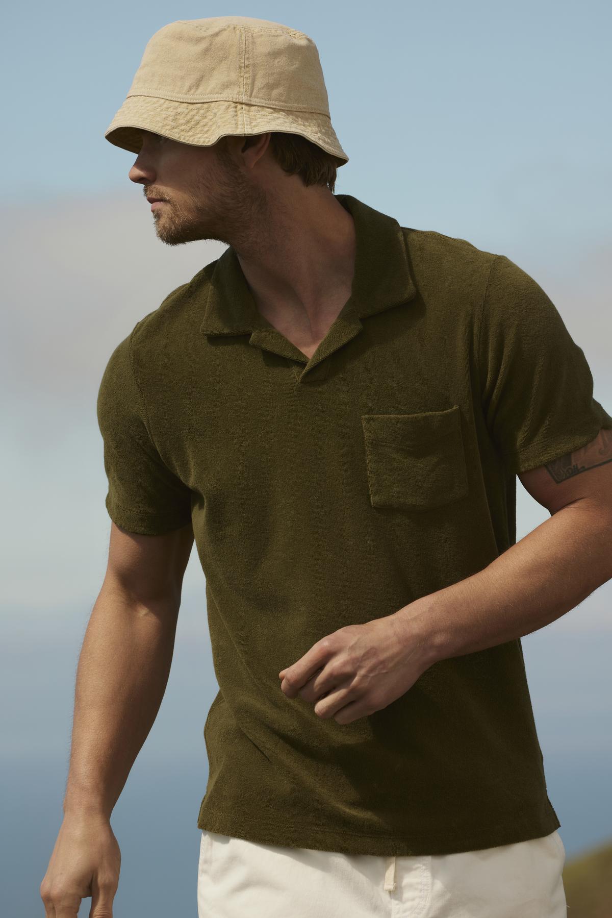 Man in a Velvet by Graham & Spencer Sergey polo shirt and beige hat, looking to the side, standing outdoors with a blurred landscape in the background.-36753587929281
