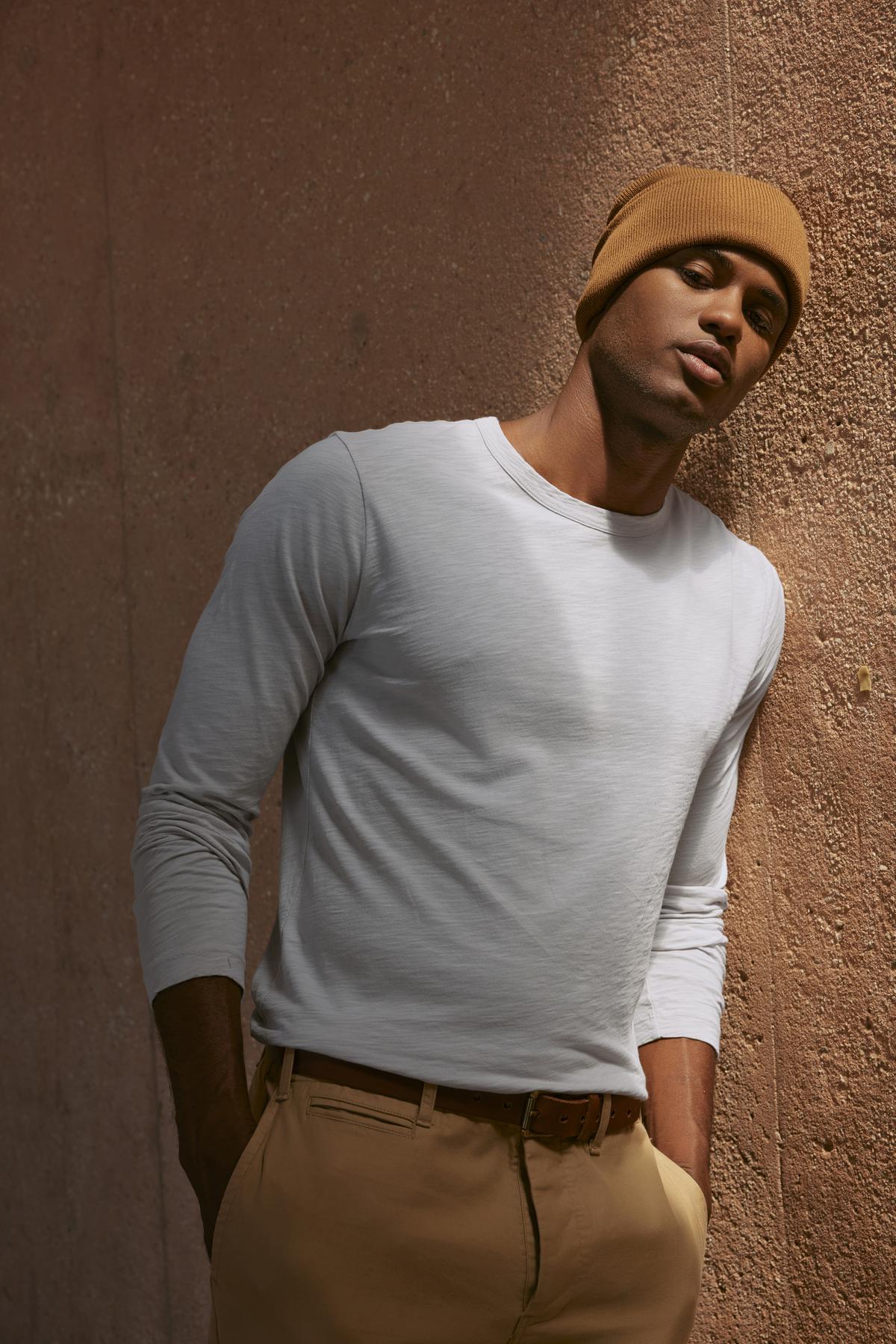   A man in the KAI TEE by Velvet by Graham & Spencer, a white long-sleeve shirt crafted from lightweight Peruvian cotton, and a brown beanie leans against a textured wall, showcasing timeless design. 