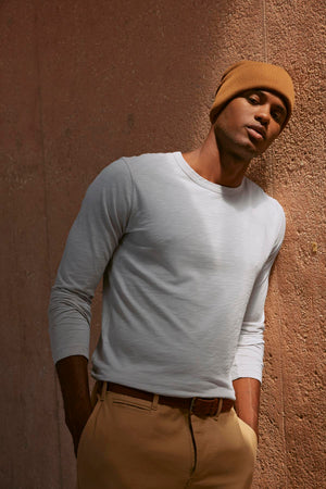 A man in the KAI TEE by Velvet by Graham & Spencer, a white long-sleeve shirt crafted from lightweight Peruvian cotton, and a brown beanie leans against a textured wall, showcasing timeless design.