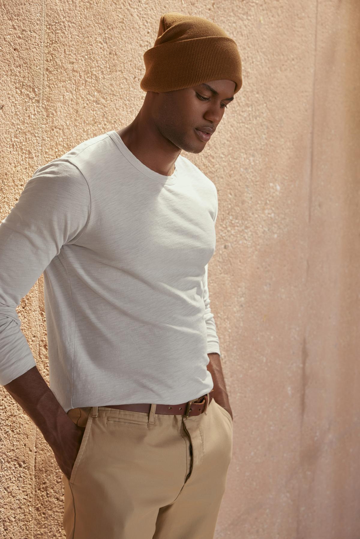   A person dressed in the KAI TEE by Velvet by Graham & Spencer, along with beige pants and a brown beanie, stands against a textured wall, highlighting a timeless design. 