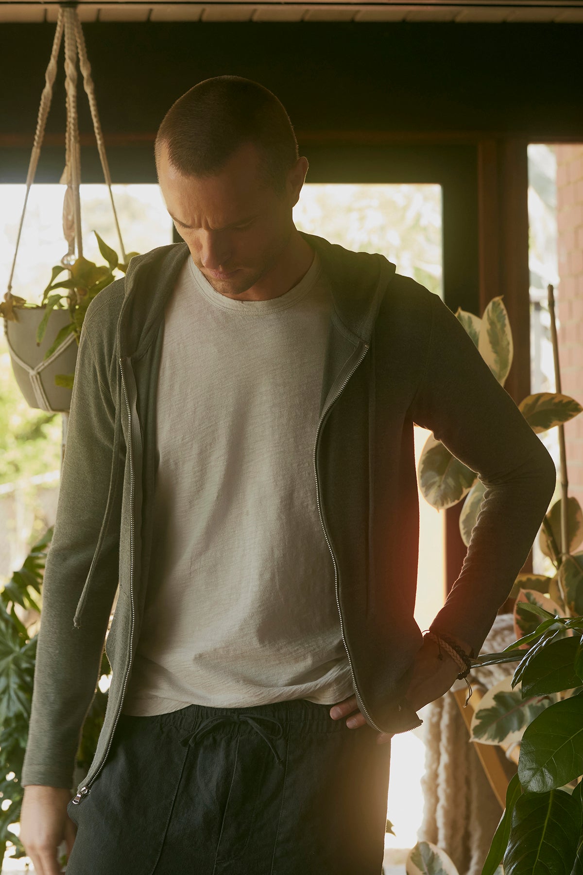 A man stands indoors near plants, wearing the Velvet by Graham & Spencer LOUDON HOODIE, a relaxed fit grey full-zip hoodie over a white t-shirt. With his hand on his hip, he looks down thoughtfully.-37759198560449