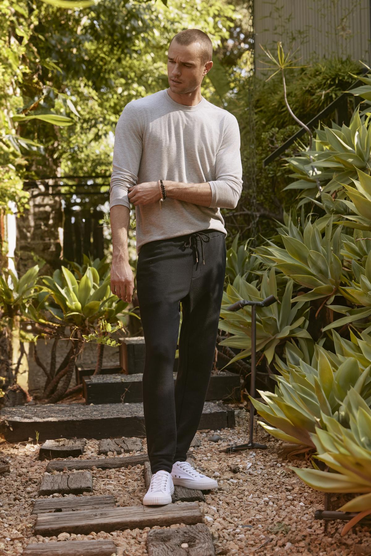   A man with a shaved head stands on a wooden path in a garden, wearing the ultra-soft BECKER CREW long-sleeve gray shirt by Velvet by Graham & Spencer, and black pants paired with white sneakers. He is looking down with one hand adjusting his sleeve. 