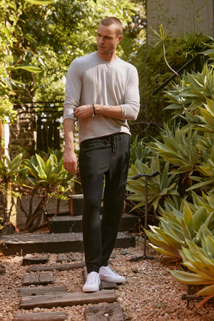 A man with a shaved head stands on a wooden path in a garden, wearing the ultra-soft BECKER CREW long-sleeve gray shirt by Velvet by Graham & Spencer, and black pants paired with white sneakers. He is looking down with one hand adjusting his sleeve.