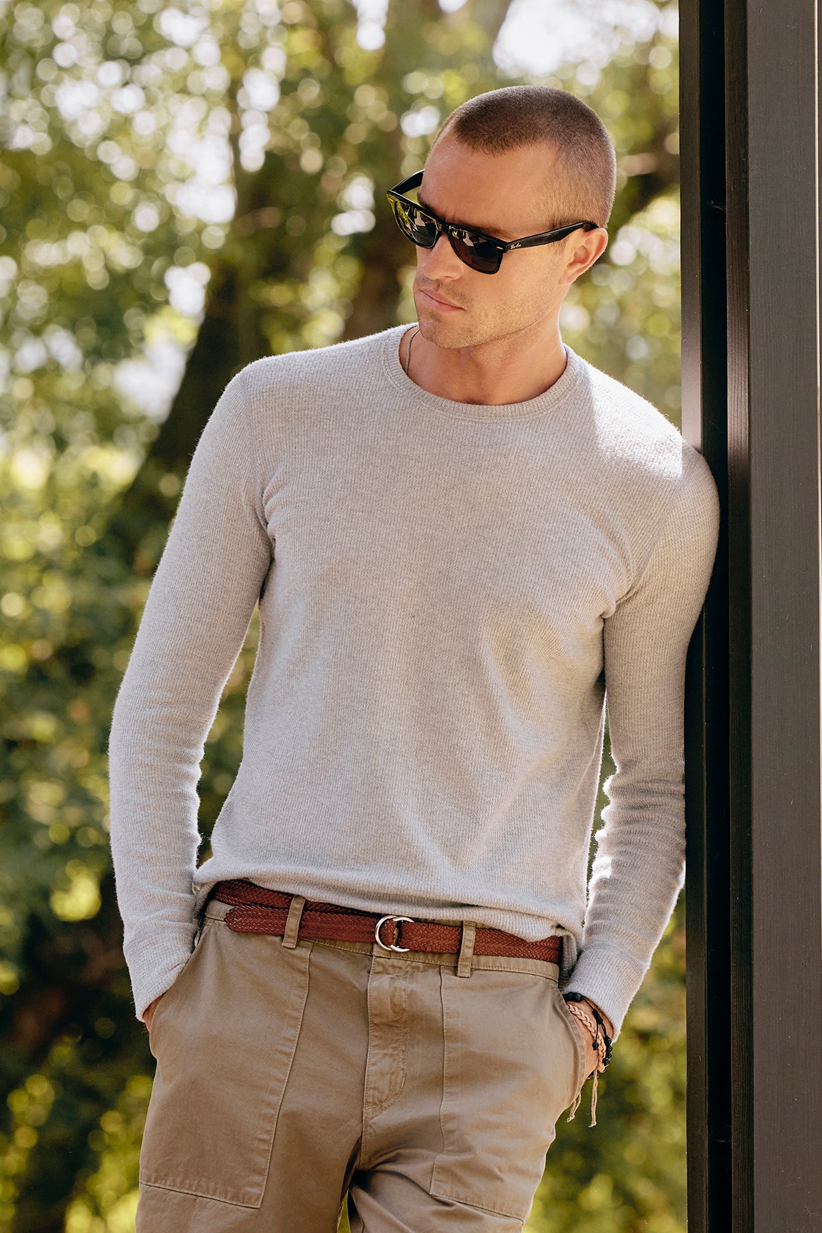   A man in sunglasses and a light sweater leans against a post, perfectly showcasing the BECKER CREW by Velvet by Graham & Spencer. The fitted crew neck design enhances his style as he stands outdoors, with trees providing a picturesque backdrop. 