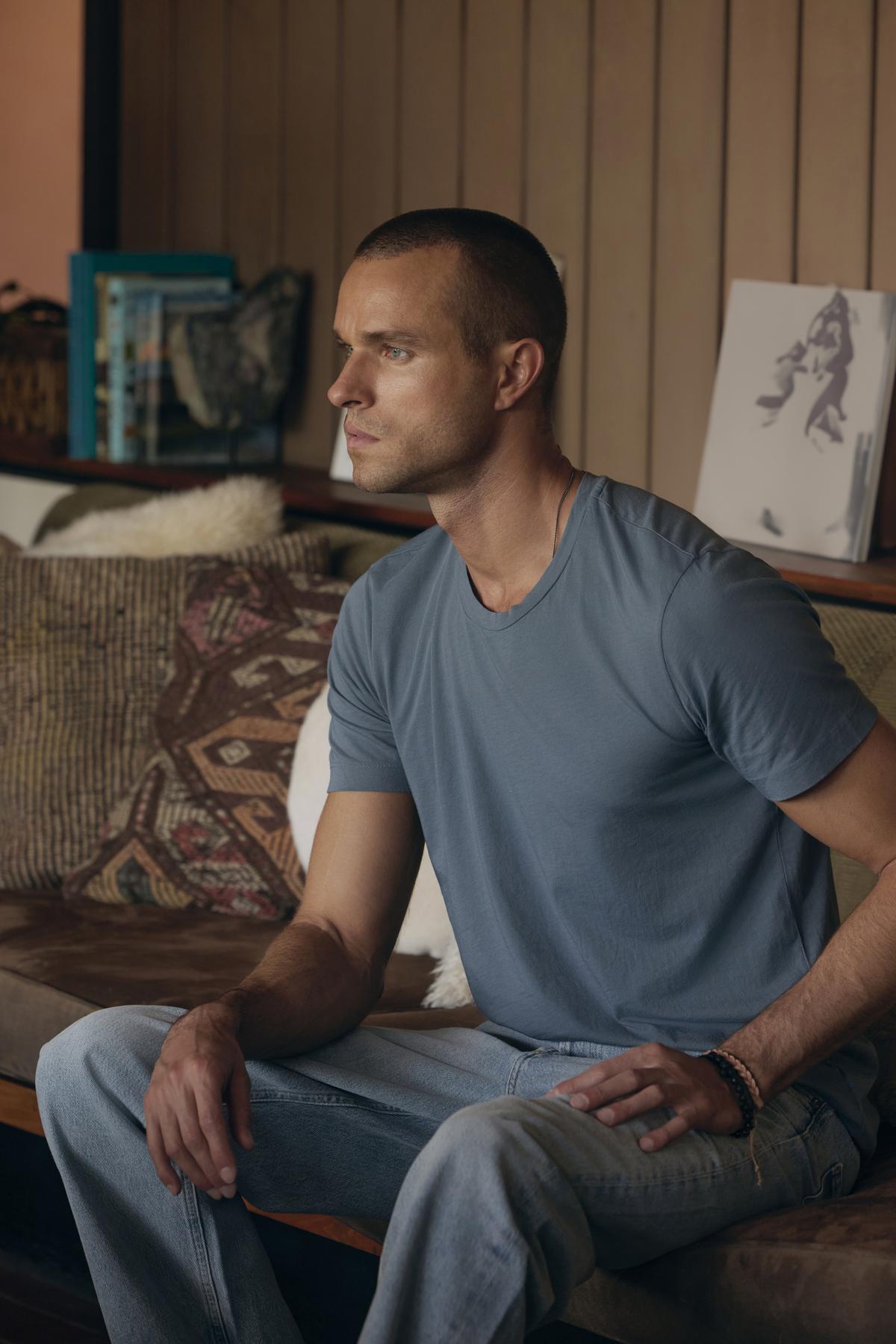 A man with short hair sits on a couch, clad in the HOWARD TEE by Velvet by Graham & Spencer, made from lightweight Pima cotton, paired with jeans. He gazes to the side with a focused expression. The background features a vintage atmosphere, complete with a wall adorned by books and framed artwork.-37759061098689