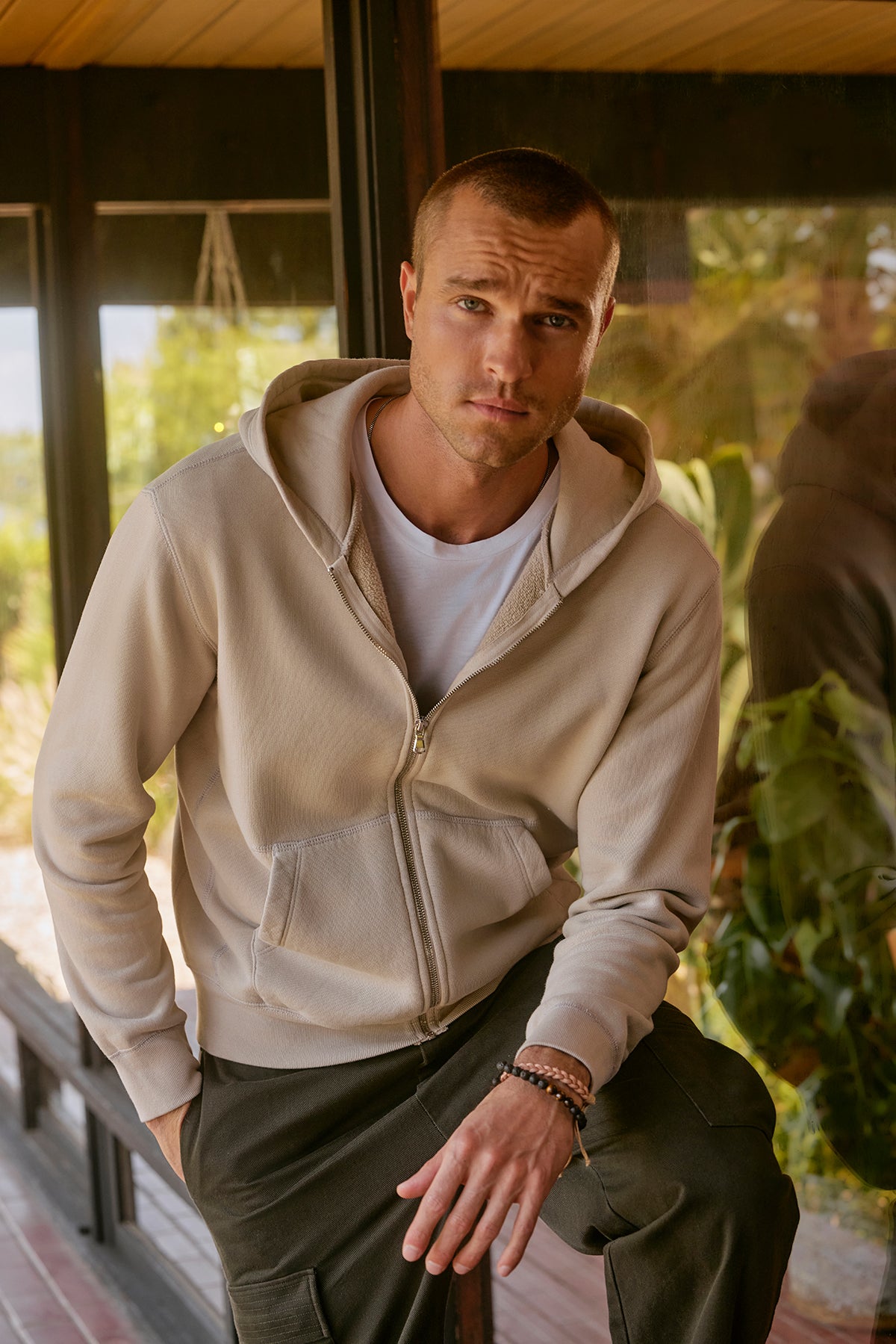   A person wearing a REN HOODIE by Velvet by Graham & Spencer along with dark pants stands indoors near a glass window, one hand casually tucked in their pocket. 