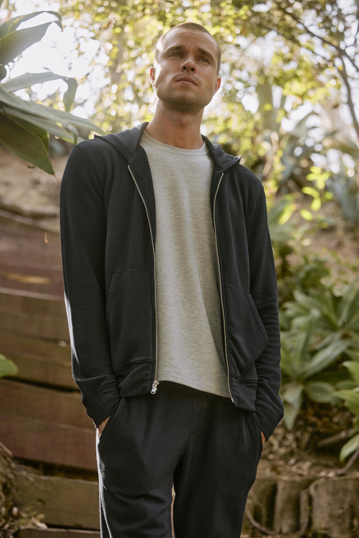 A man stands outdoors in casual attire, sporting the RODAN HOODIE by Velvet by Graham & Spencer along with pants and a light grey t-shirt underneath. He is looking upwards, surrounded by greenery and steps in the background, making his hoodie a perfect workout layer for the serene setting.-37888558629057