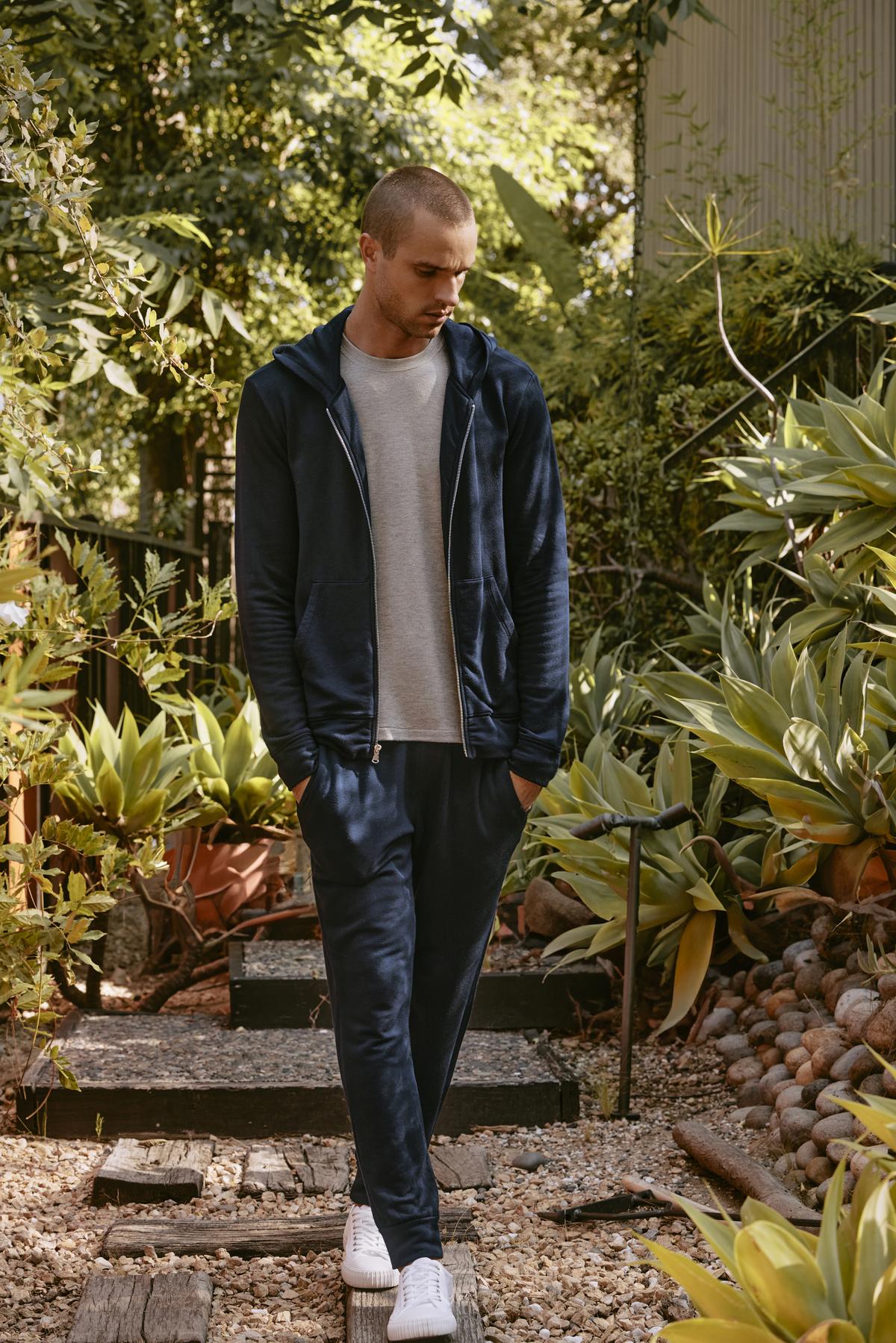   A person in a tracksuit featuring Velvet by Graham & Spencer's CROSBY JOGGER and white sneakers stands on a garden path surrounded by lush green plants. 