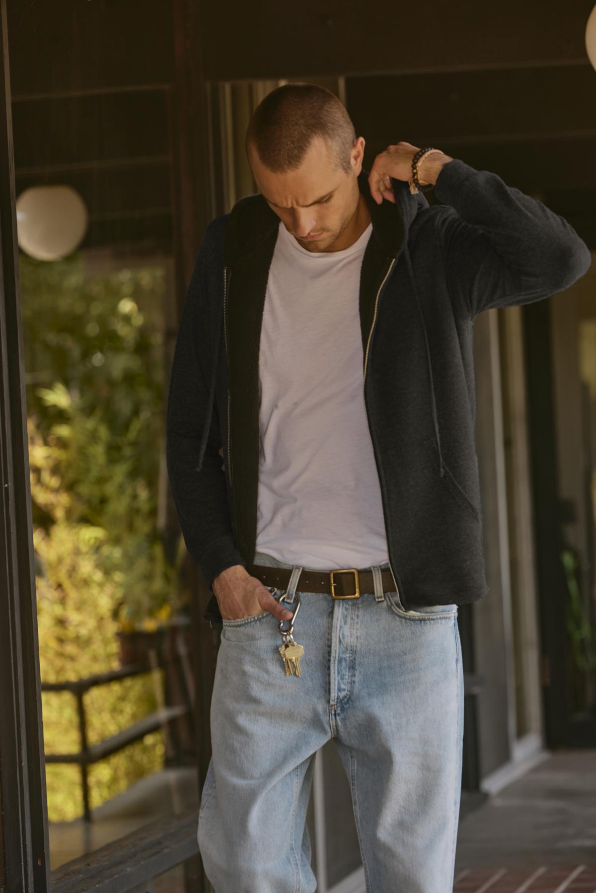   A person with a shaved head, wearing the ultra-soft SALVADORE HOODIE by Velvet by Graham & Spencer, a white t-shirt, and light blue jeans, stands outside looking down while adjusting their belt. Keys are attached to the belt loop on their jeans. 