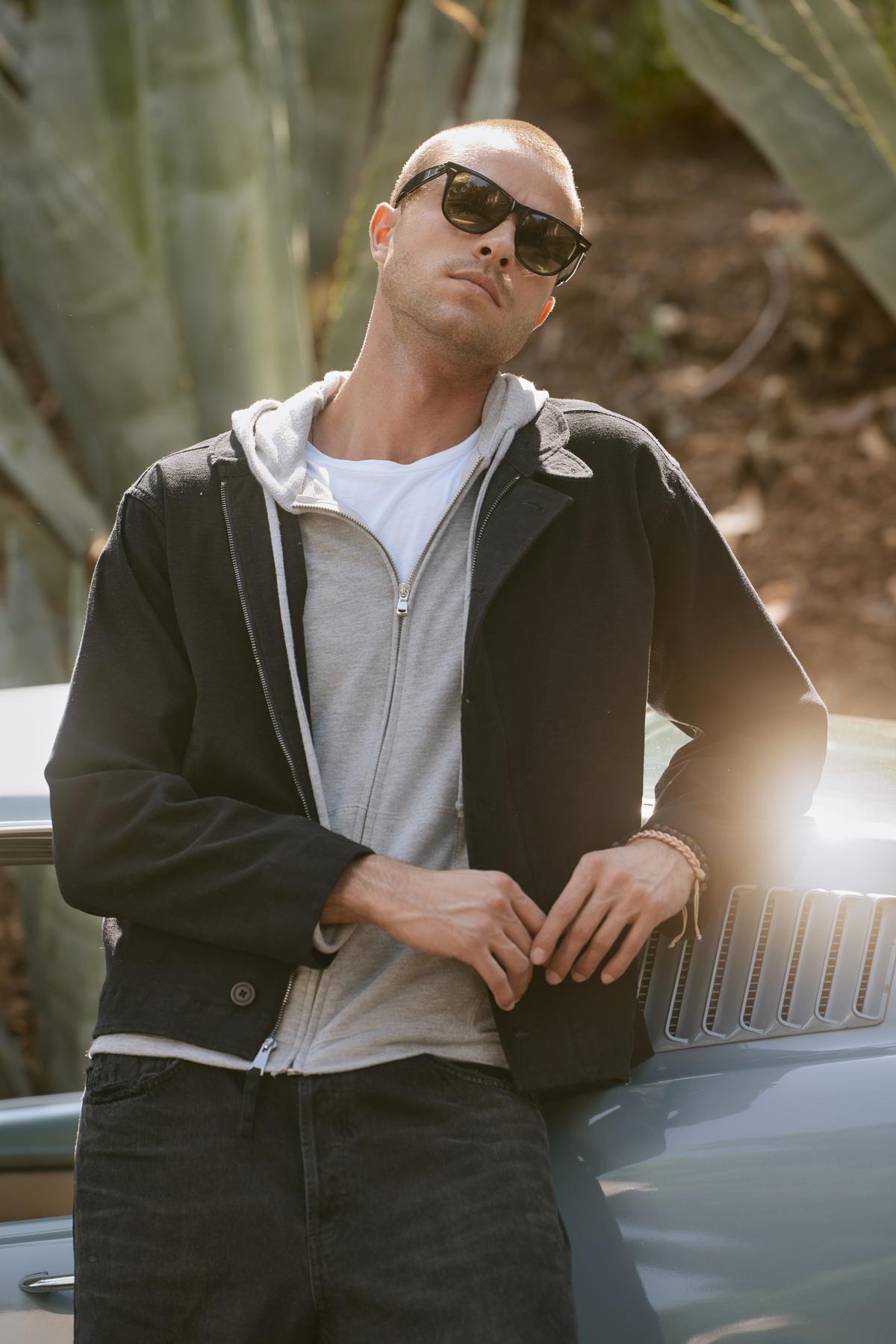 A person wearing sunglasses, a black jacket, and the LOUDON HOODIE by Velvet by Graham & Spencer leans against a car in an outdoor setting.-37888579207361