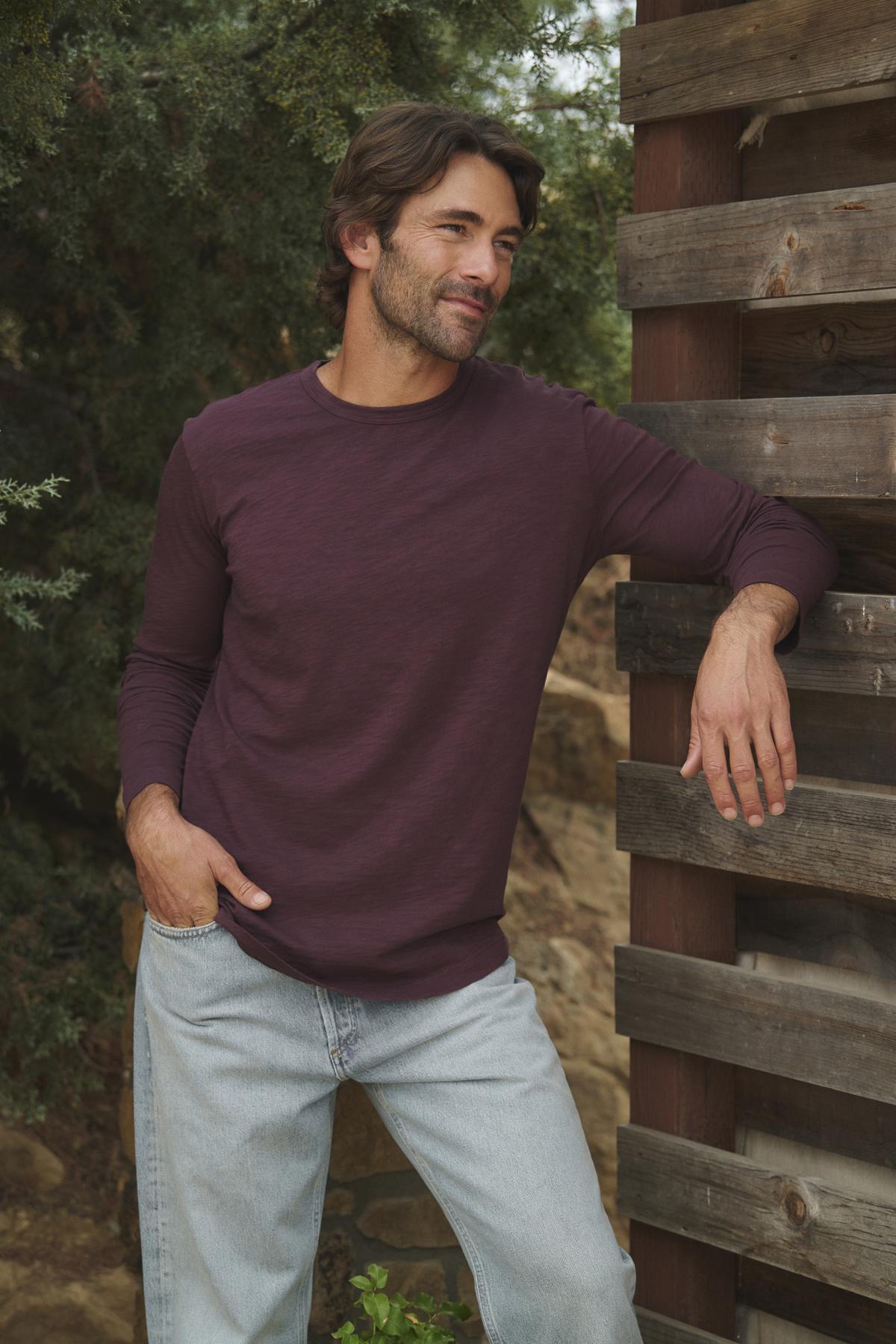   A man wearing the KAI TEE by Velvet by Graham & Spencer, a maroon long-sleeve shirt featuring a classic crew neckline, pairs it with light jeans as he leans against a wooden structure outdoors amidst lush greenery. The timeless design of his outfit highlights the elegance of lightweight Peruvian cotton. 