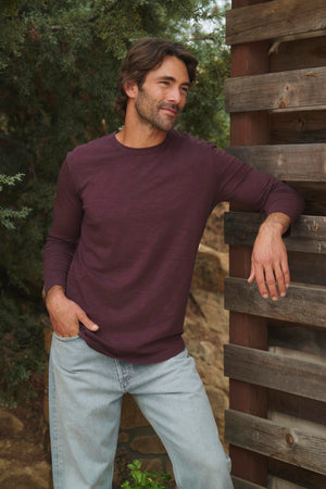 A man wearing the KAI TEE by Velvet by Graham & Spencer, a maroon long-sleeve shirt featuring a classic crew neckline, pairs it with light jeans as he leans against a wooden structure outdoors amidst lush greenery. The timeless design of his outfit highlights the elegance of lightweight Peruvian cotton.