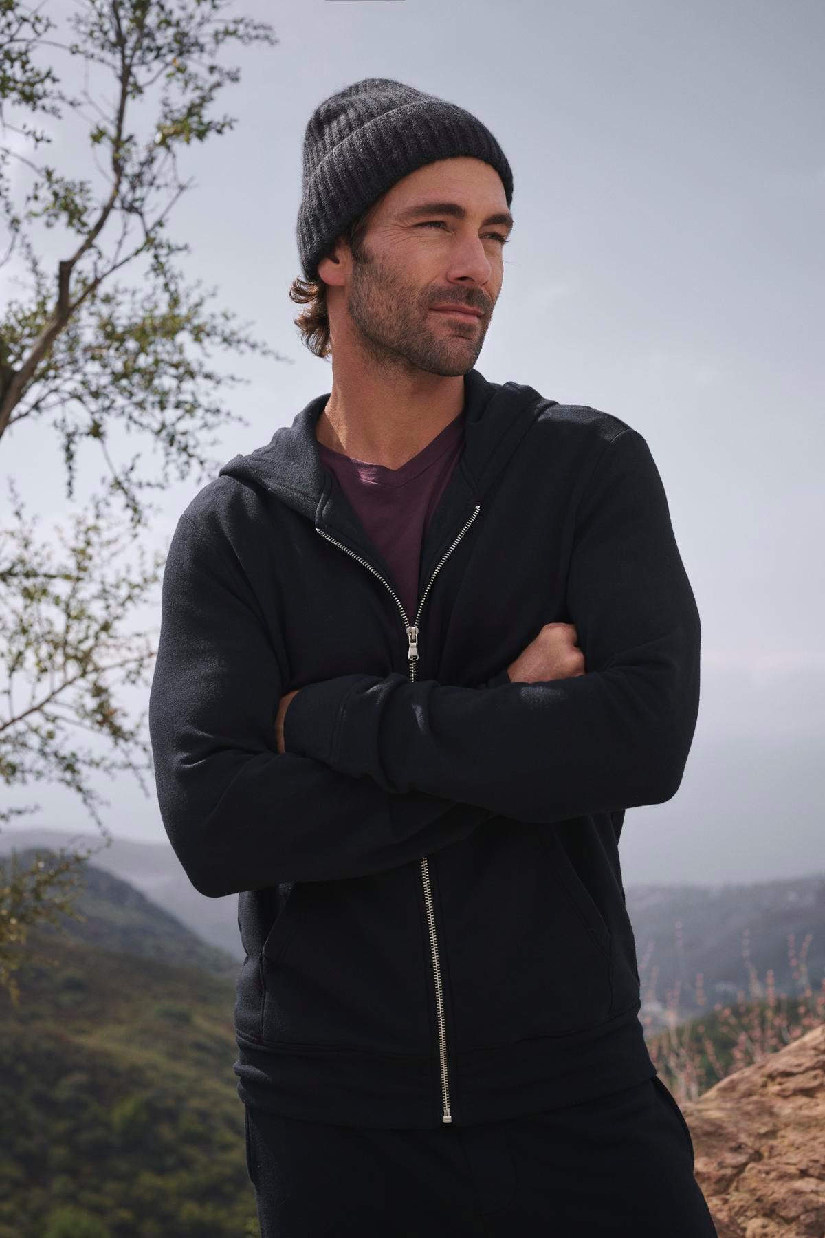   A person is standing outdoors, with arms crossed and facing the camera, wearing the RODAN HOODIE by Velvet by Graham & Spencer. The backdrop consists of trees and hills. 