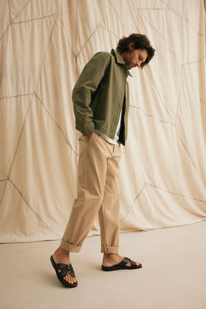 A person in a Velvet by Graham & Spencer COLLINS JACKET, beige pants, and black sandals stands against a beige, textured fabric backdrop.