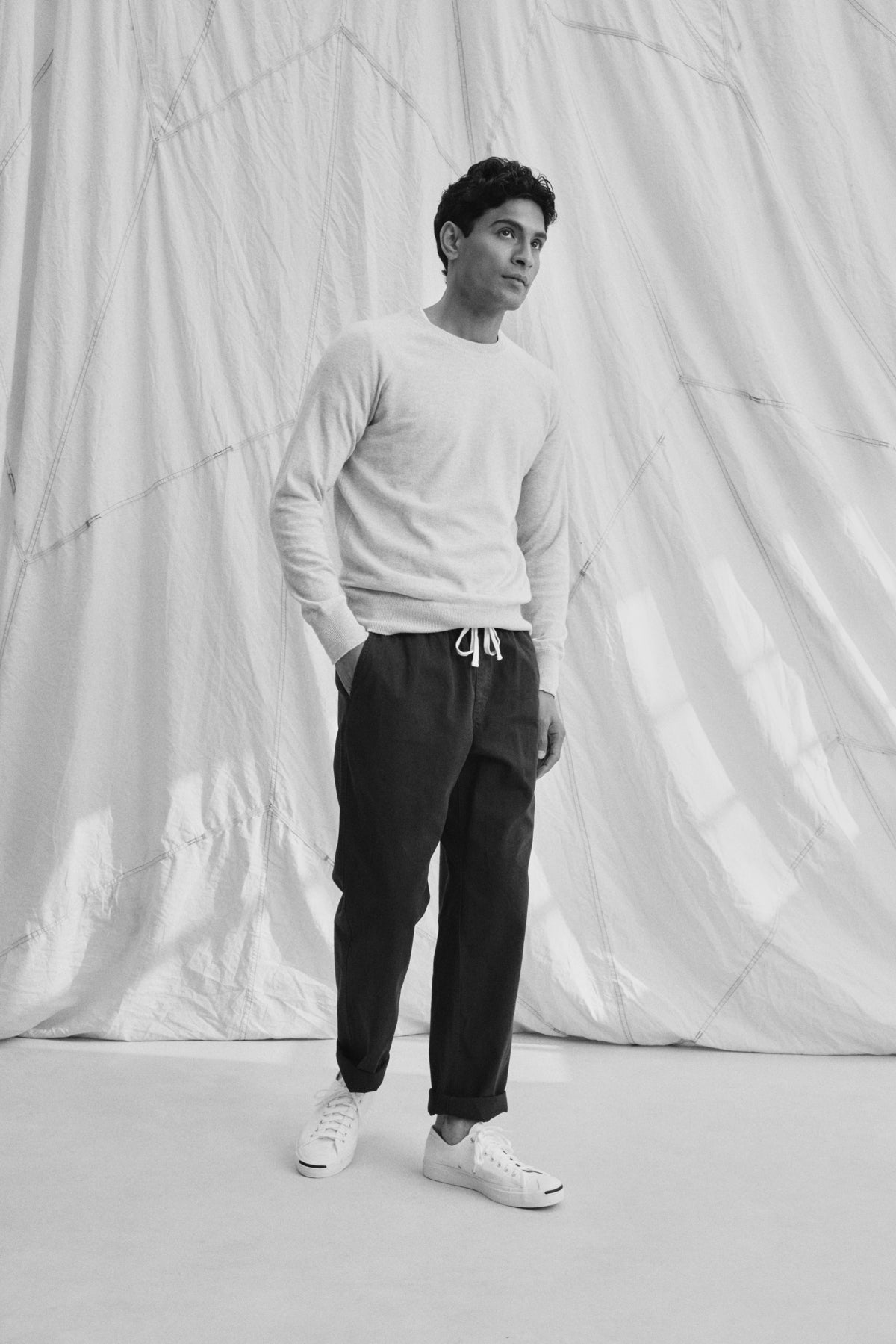   A person stands with hands in pockets, wearing a light sweater and BRANSON PANT by Velvet by Graham & Spencer. These cotton sanded twill drawstring pants feature an elastic waist and straight leg design. They sport sneakers against a draped fabric backdrop. 