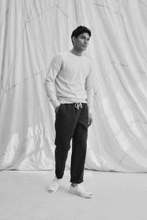 A person stands with hands in pockets, wearing a light sweater and BRANSON PANT by Velvet by Graham & Spencer. These cotton sanded twill drawstring pants feature an elastic waist and straight leg design. They sport sneakers against a draped fabric backdrop.