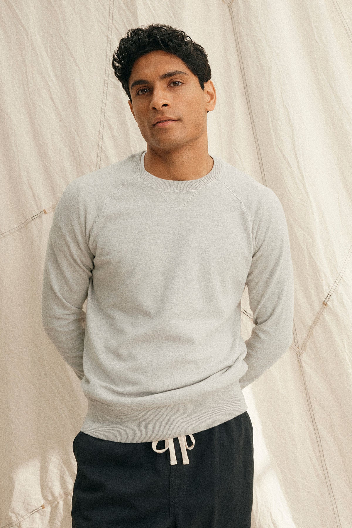   A person wearing a DARIO COTTON CASHMERE SWEATER by Velvet by Graham & Spencer in light gray and dark pants stands with hands behind their back against a beige fabric backdrop. 