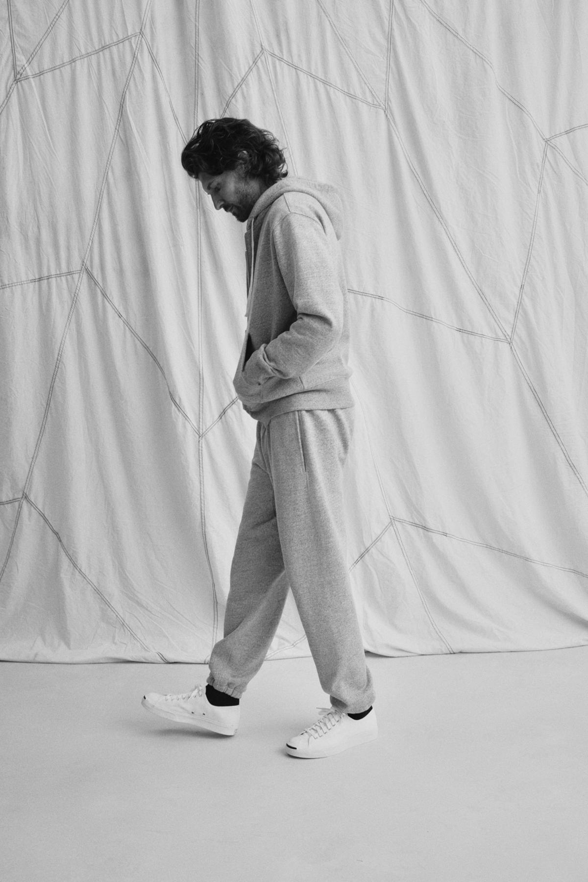 A man wearing a DRACO HOODIE by Velvet by Graham & Spencer and white sneakers walks past a white, textured curtain.-38643384123585