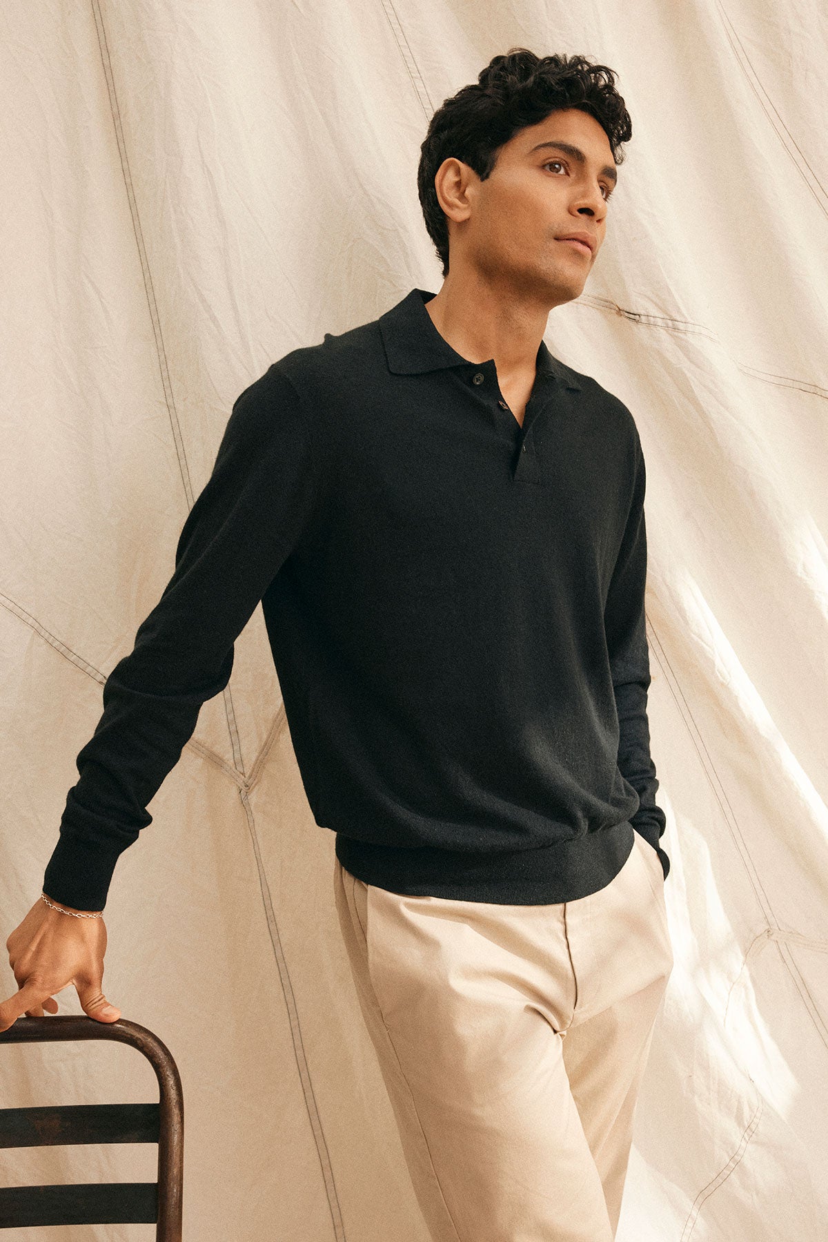   Wearing the MATTEO COTTON LINEN SWEATER POLO by Velvet by Graham & Spencer, this individual in a black long-sleeve shirt and beige pants leans against a chair, gazing upward. A draped white fabric backdrop accentuates the outfit's lightweight comfort and style. 