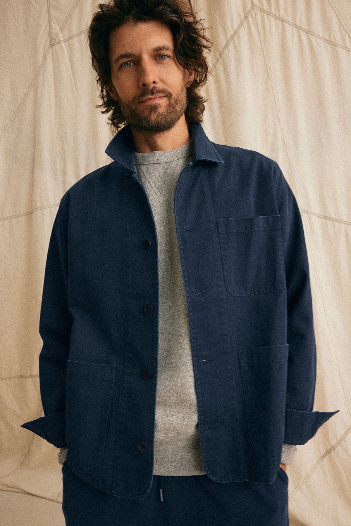   A bearded man with curly hair wears a Velvet by Graham & Spencer STEEL CHORE JACKET in navy blue over a gray sweater, standing against a light fabric backdrop. 