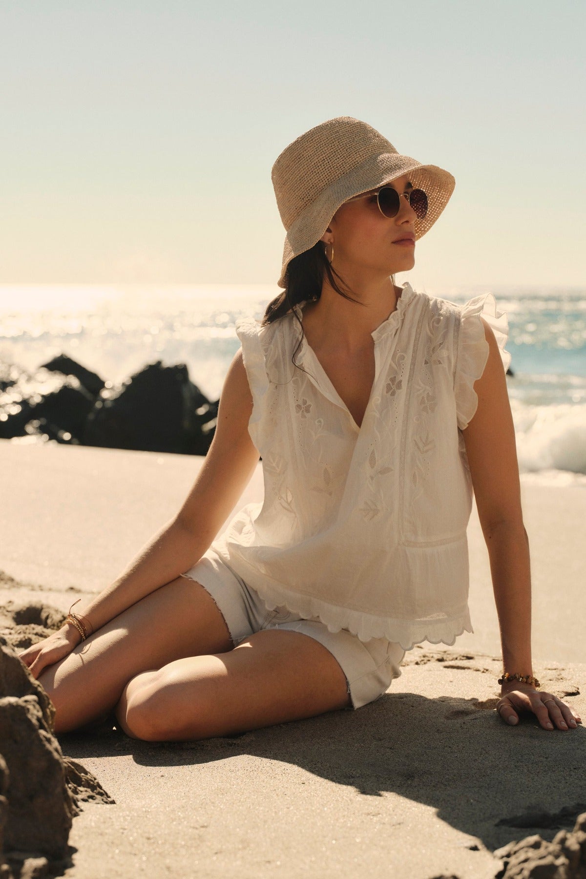 Woman in a white CHARLENE TOP by Velvet by Graham & Spencer embroidery outfit and sunhat sitting on a beach with rocks, looking away from the camera, ocean in the background.-36752743465153
