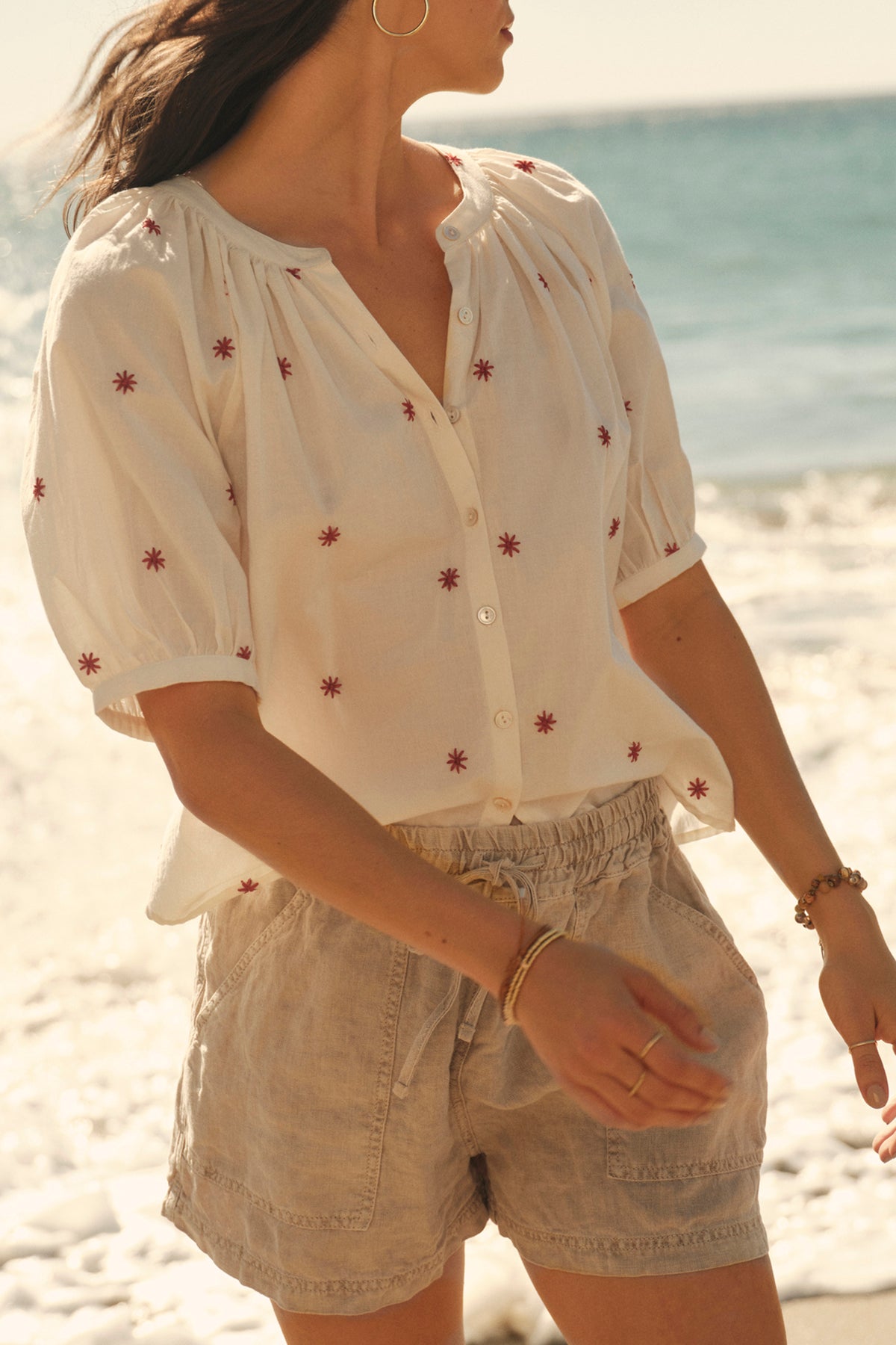Woman in a white blouse with red floral embroidery, featuring a split neck, and beige shorts, walking on a sunny beach wearing the AMIRA TOP by Velvet by Graham & Spencer.-36910000439489