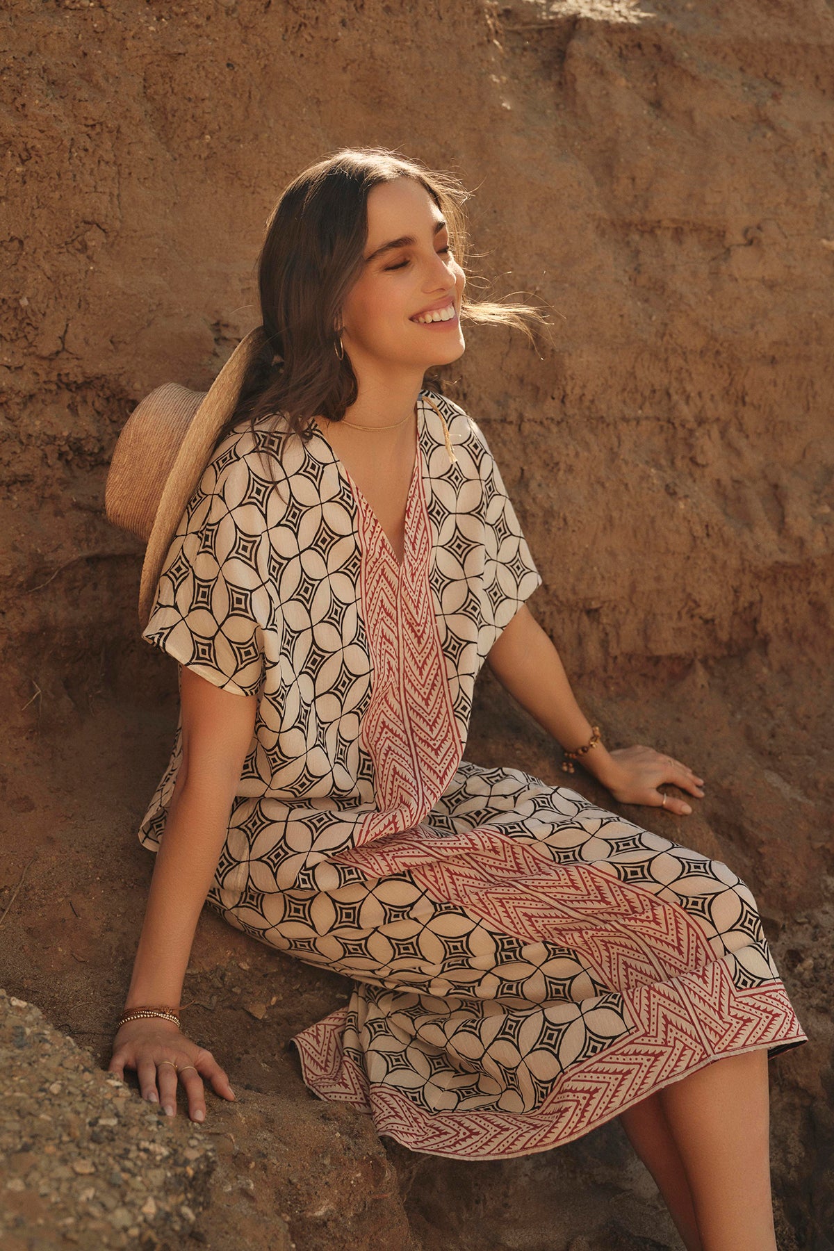   A woman in a lightweight cotton Velvet by Graham & Spencer Odessa kaftan dress smiles while sitting on a rocky terrain, enjoying the sunlight. 