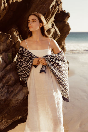 A woman in a white dress and patterned shawl from Velvet by Graham & Spencer stands beside a rocky beach, gazing to her left with the ocean in the background.