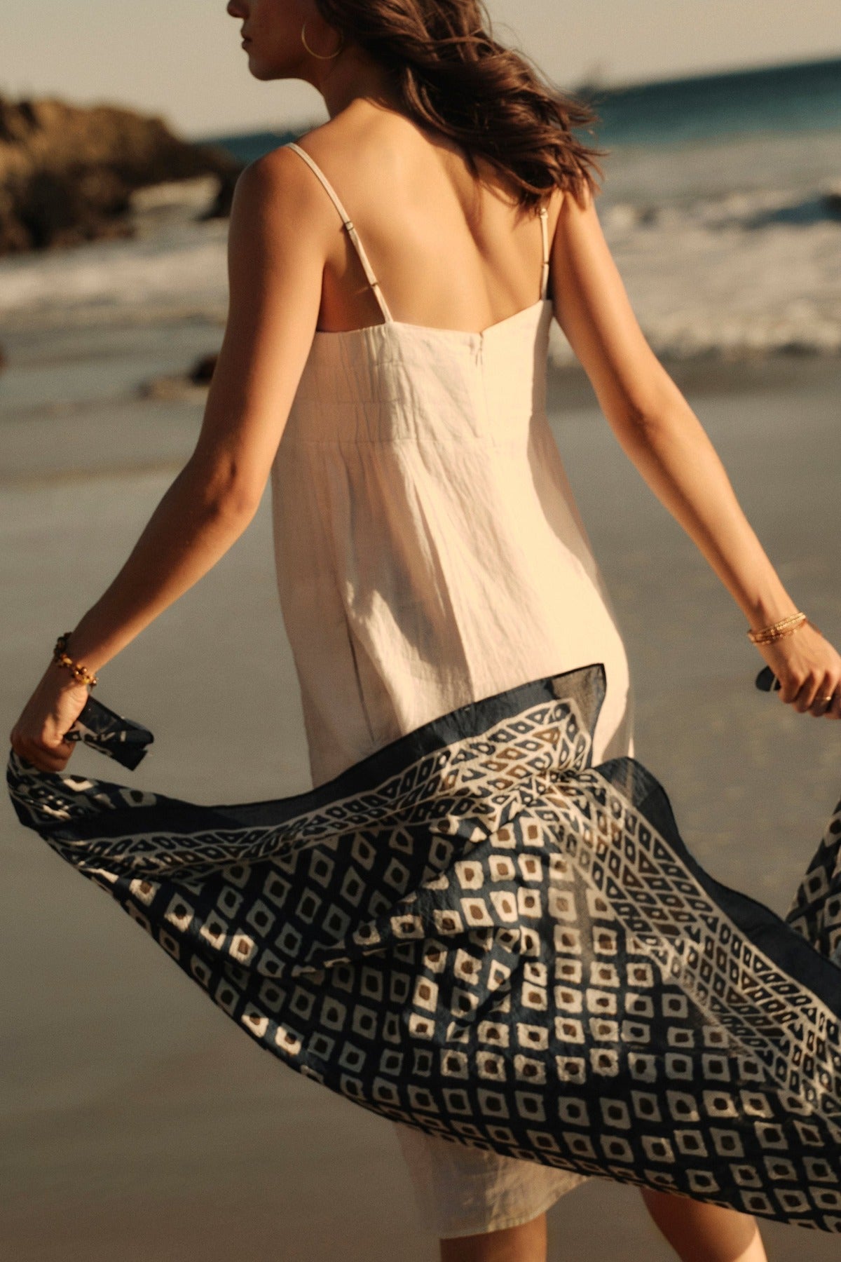   A woman in a white dress and holding a Velvet by Graham & Spencer sarong wrap walks along a sandy beach at sunset. 