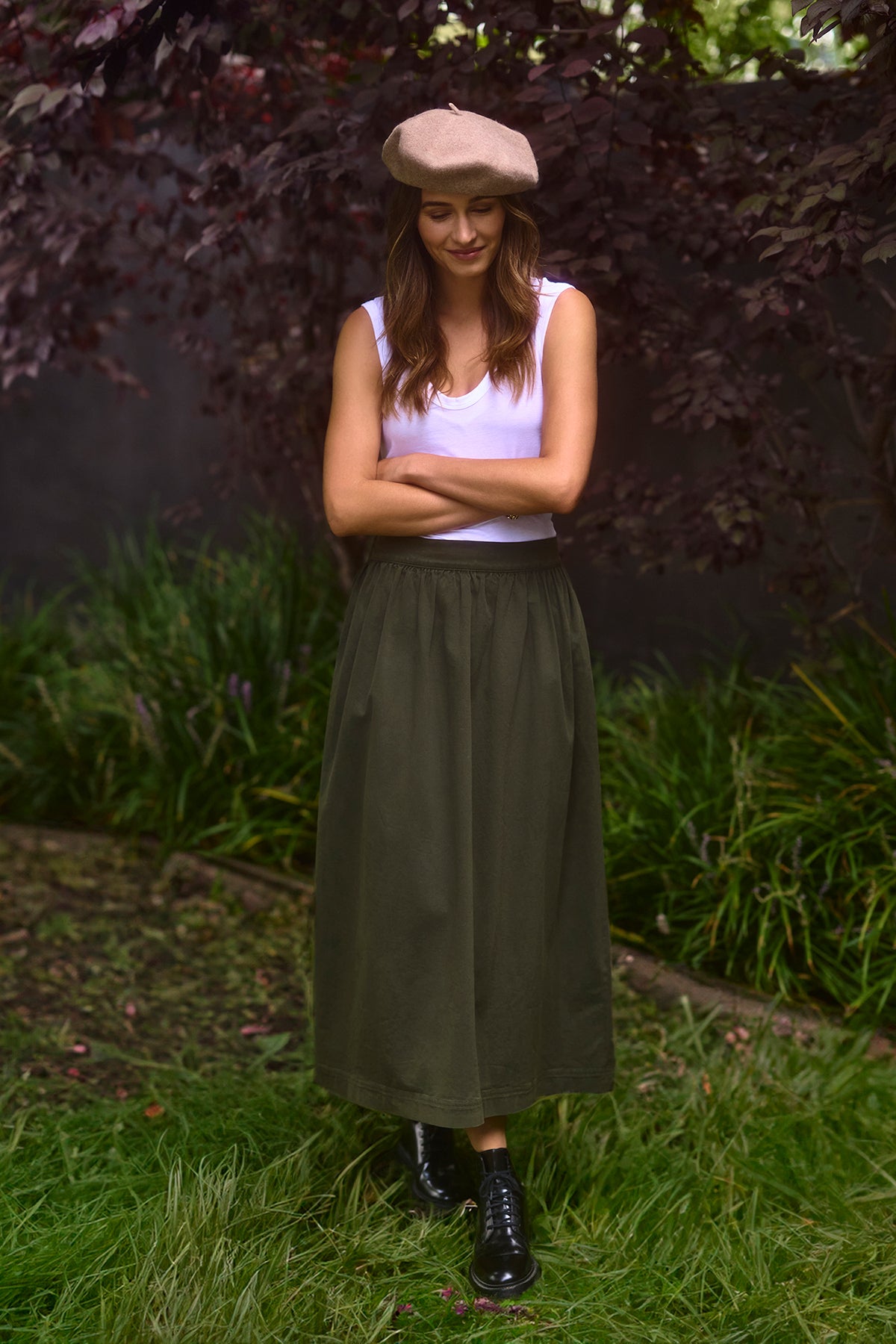   A woman wearing a white tank top, black boots, and a beige beret stands on a grassy area with her arms folded, looking down and smiling. Her Velvet by Graham & Spencer SOPHIA SKIRT in olive green stretch cotton twill gracefully complements her relaxed yet stylish demeanor. 