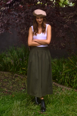 A woman wearing a white tank top, black boots, and a beige beret stands on a grassy area with her arms folded, looking down and smiling. Her Velvet by Graham & Spencer SOPHIA SKIRT in olive green stretch cotton twill gracefully complements her relaxed yet stylish demeanor.