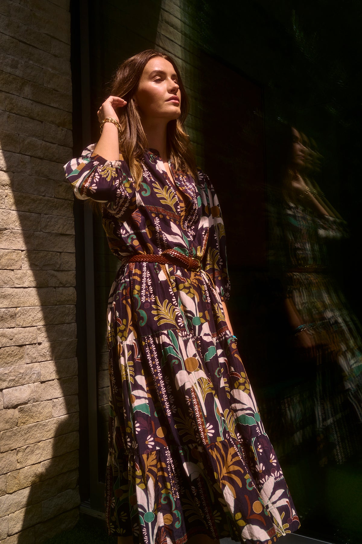   A woman in the JOHANNA PRINTED SILK COTTON VOILE DRESS from Velvet by Graham & Spencer, featuring a bold, floral-patterned midi-length skirt, stands beside a wall with her hand touching her hair. She casts a shadow, and her reflection is visible on the glass surface next to her. 